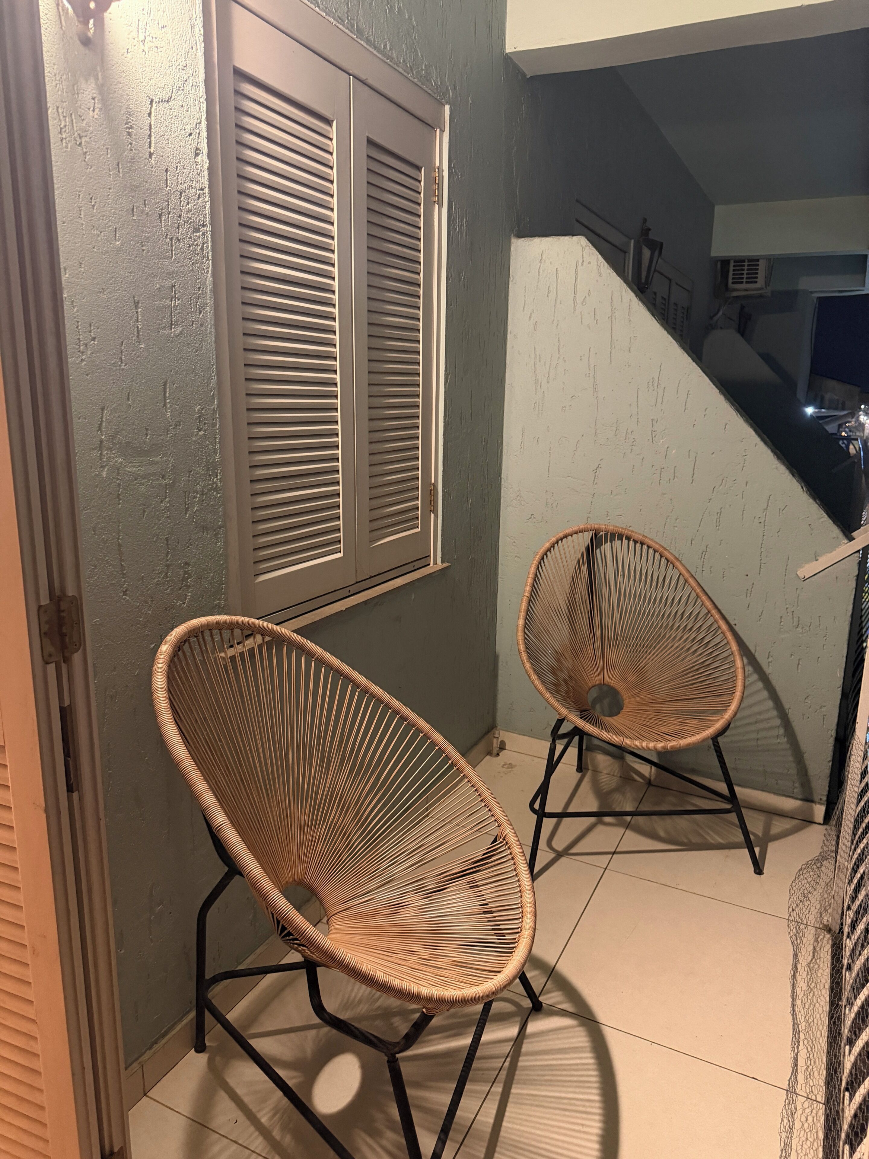 A room with two wicker chairs, a window with shutters, and a staircase leading to an upper level.