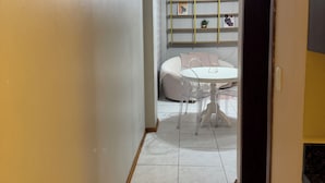 A hallway leading to a dining area with a round table and chairs, a sofa, and a wall-mounted shelf with decorative items.