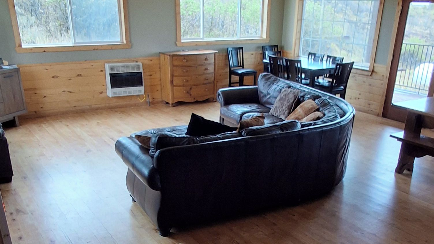 Una sala de estar acogedora con un sofá negro curvo, suelo de madera, un mueble pequeño de madera y un comedor con sillas y una mesa.