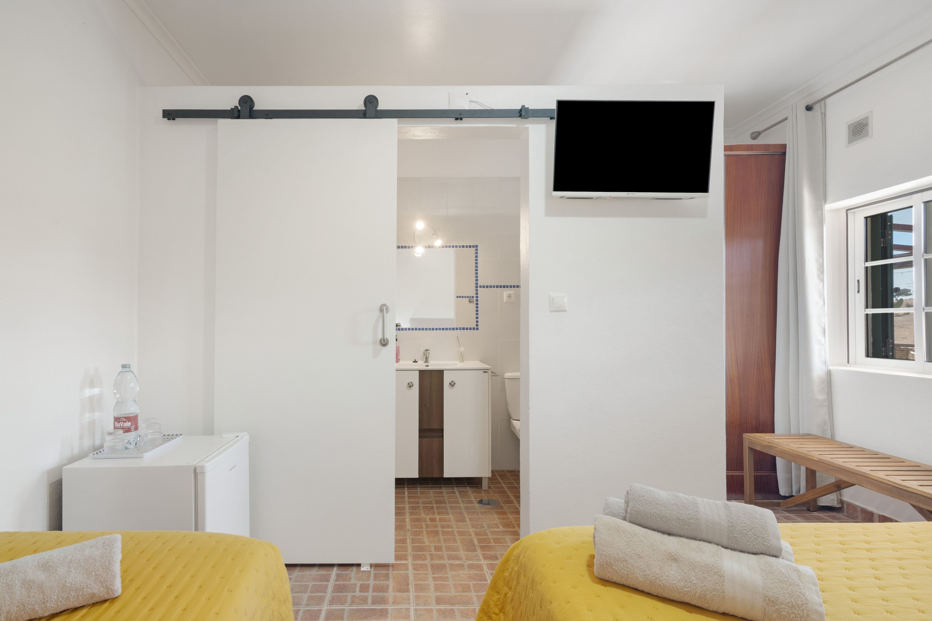 Ein Hotelzimmer mit zwei Betten, einem Fernseher, einer Holzbank und einem durch eine offene Tür einsehbaren Badezimmer.