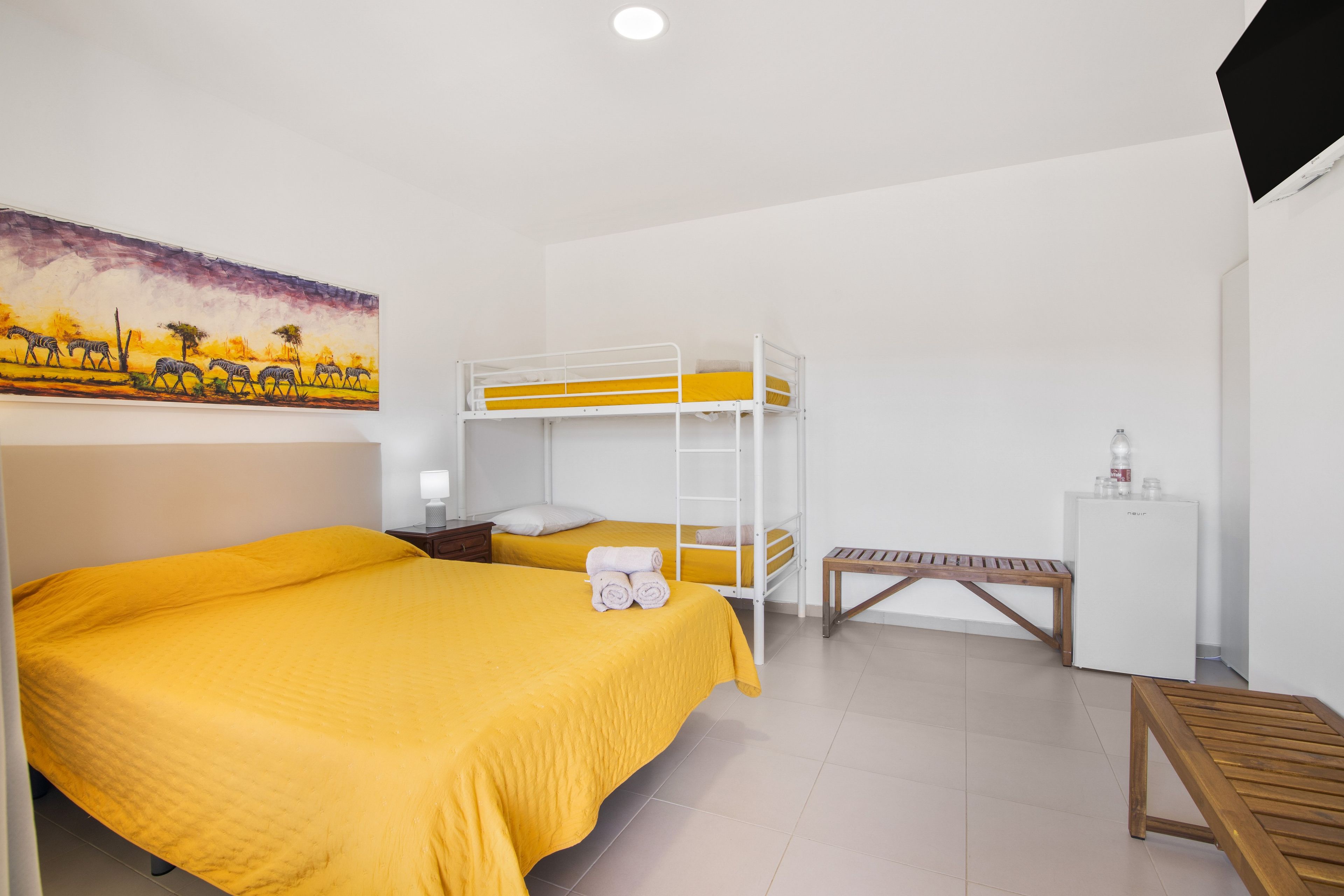 Una habitación con dos camas, un banco, una mesita y un cuadro en la pared.