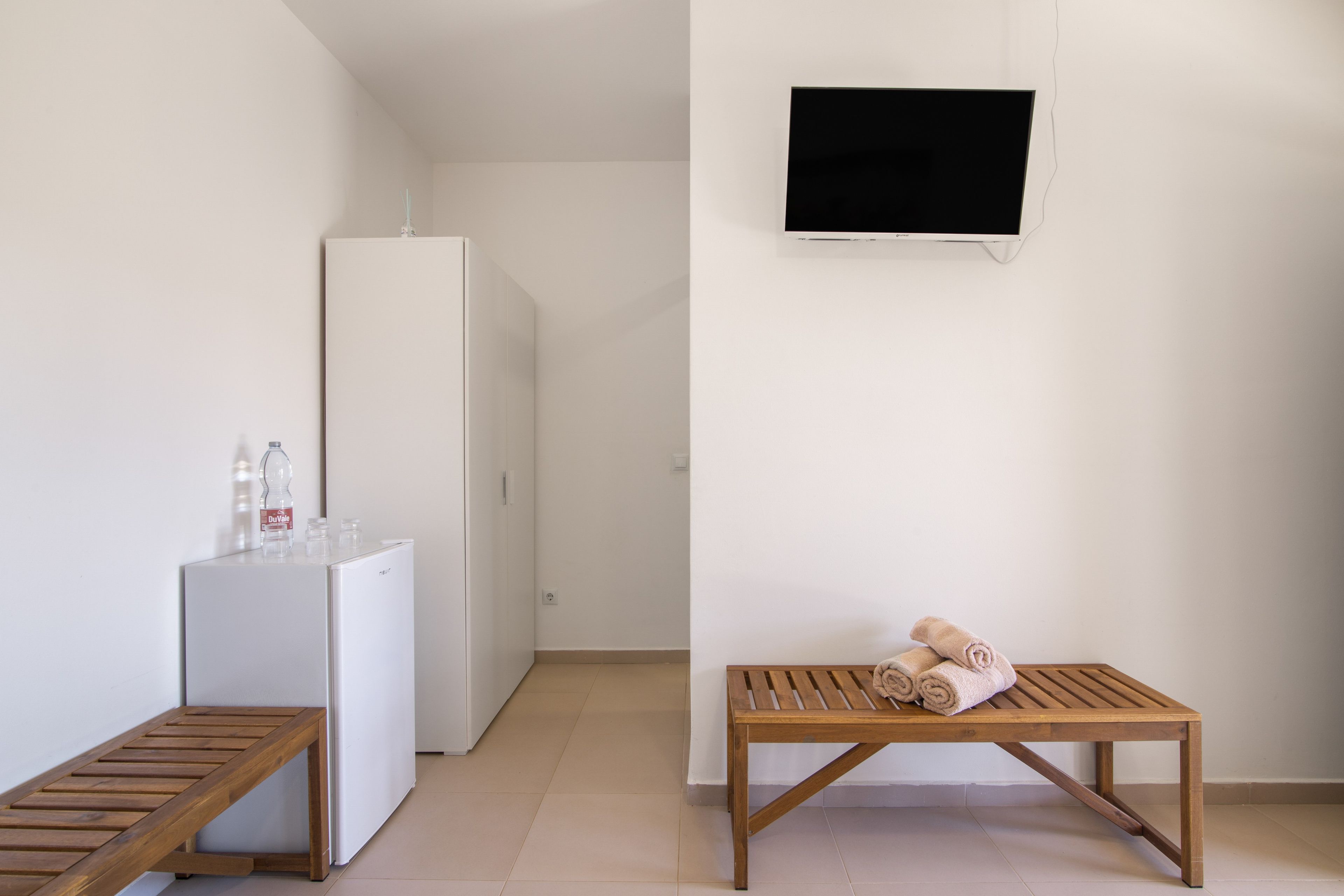 Ein schlicht eingerichteter Raum mit einem weißen Kühlschrank, einer Holzbank, einem kleinen Tisch mit Handtüchern und einem Wand-Fernseher.