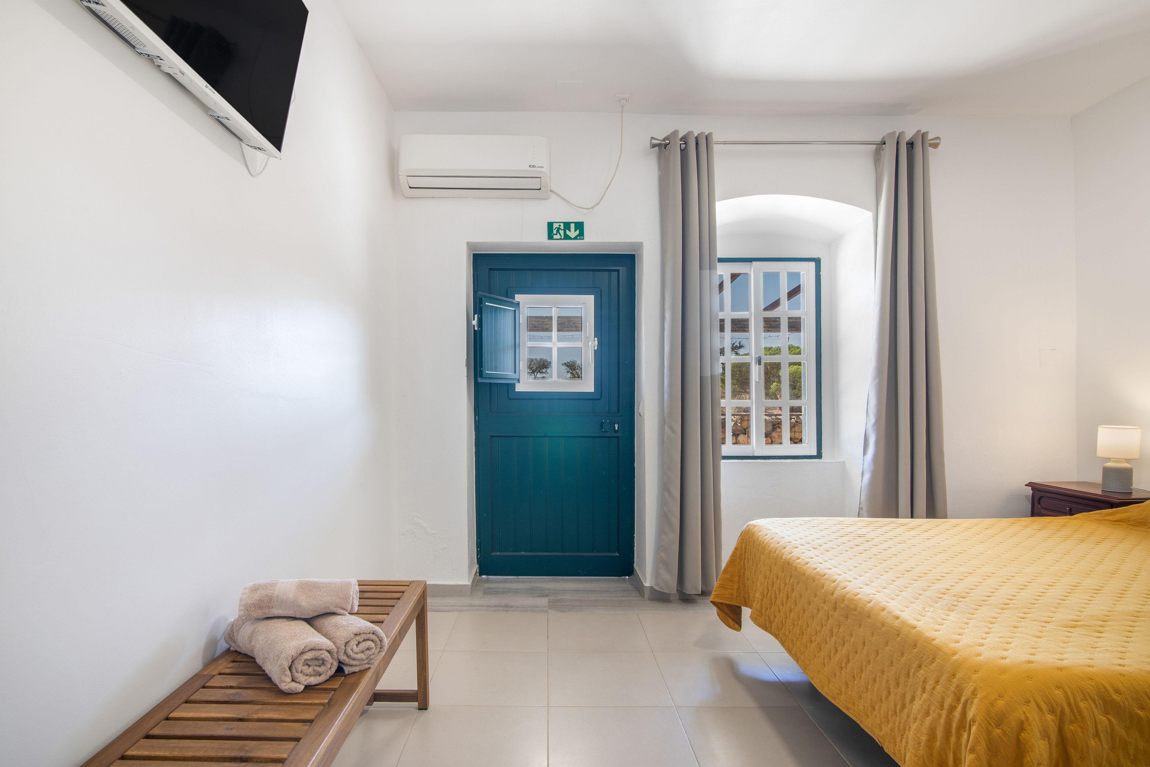 A hotel room with a bed, a bench with towels, a door, and a window.