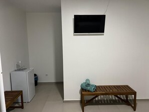 Una habitación con un banco de madera, una mesa pequeña, un refrigerador blanco y una televisión instalada en la pared.