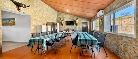 A dining area with a long table set for multiple people, stone walls, a wooden ceiling, and a view through large windows.