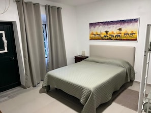 Een slaapkamer met een bed, gordijnen, een schilderij, een nachtkastje en een deur.