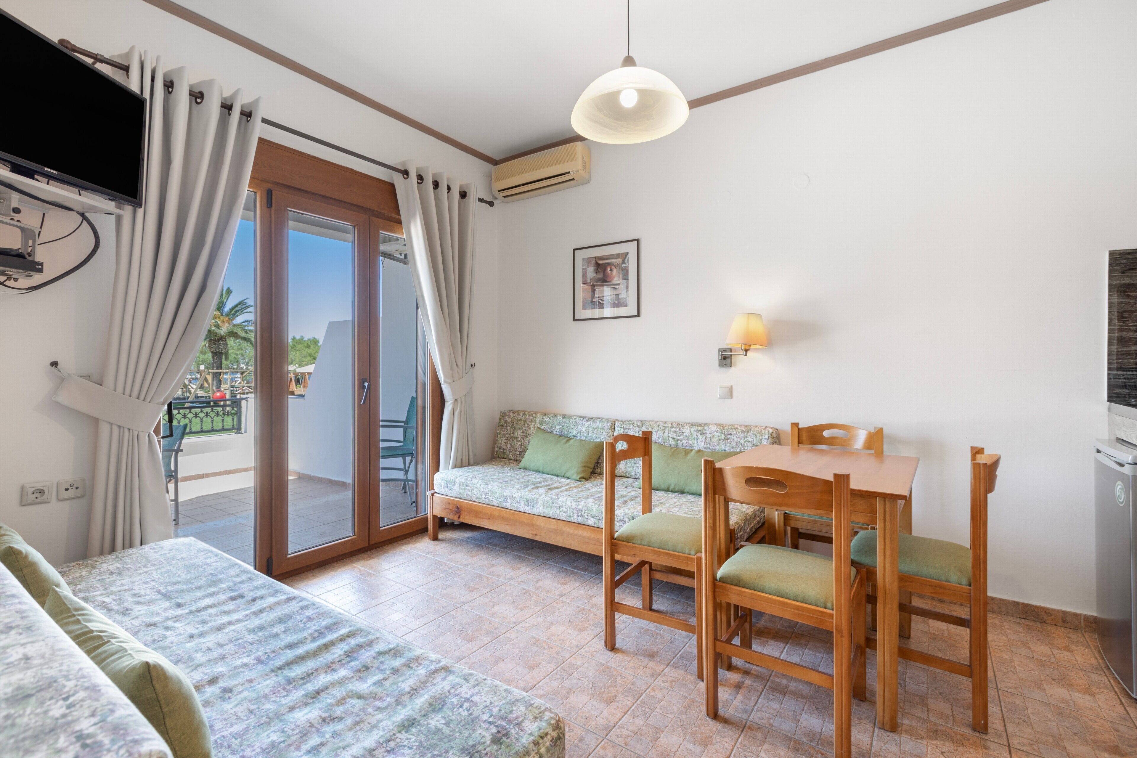 Une pièce comprenant un canapé, une table à manger avec des chaises, un téléviseur fixé au mur et une porte-fenêtre coulissante donnant sur un espace extérieur.
