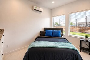 Une chambre à coucher avec un lit, une fenêtre avec des stores, un appareil de climatisation et une petite table sur laquelle est posée une plante.