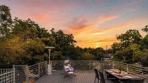 En træterrasse med spisebord og stole, omgivet af træer og en solnedgang.