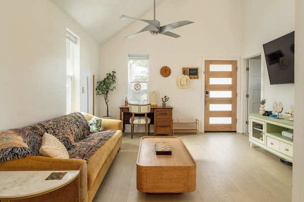 A living room with a sofa, coffee table, and a desk with a chair. There is a ceiling fan, a door with frosted glass panels, and a wall-mounted TV. - La Holly Rooftop Retreat (Austin)
