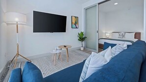 Et moderne hotelværelse med et fladskærms-tv, en seng med blåt sengetøj, et lille bord med en lampe og en potteplante.