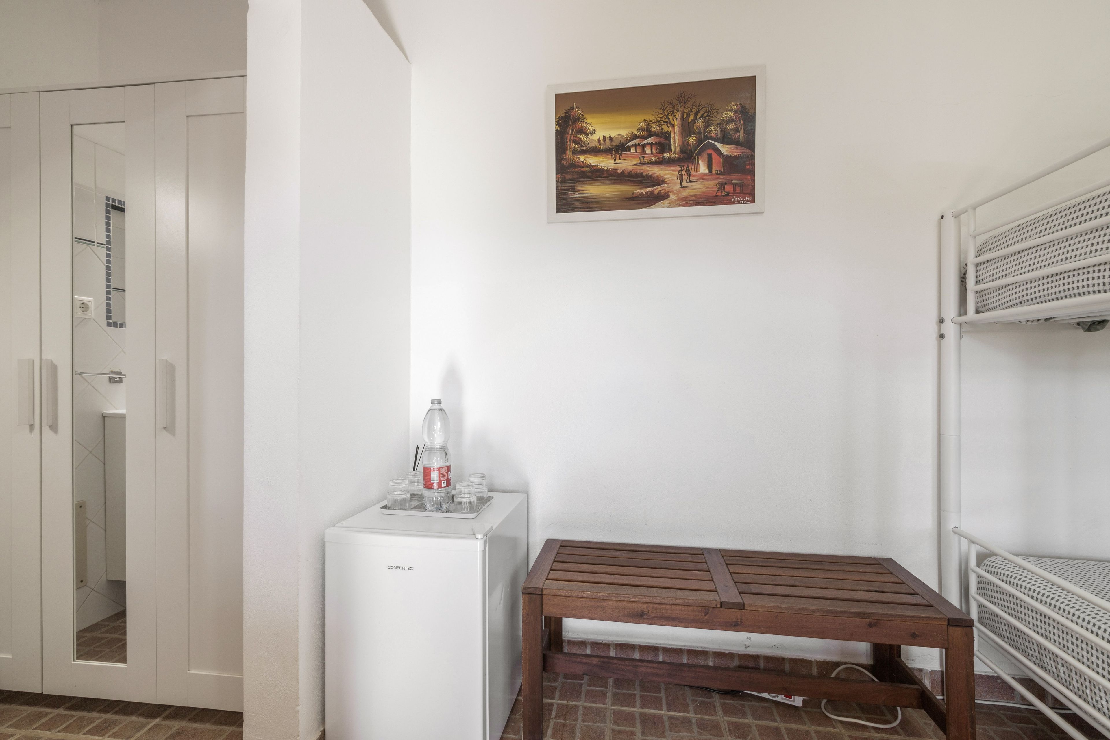 A compact room with a white door, a small refrigerator, a wooden bench, and a bunk bed.