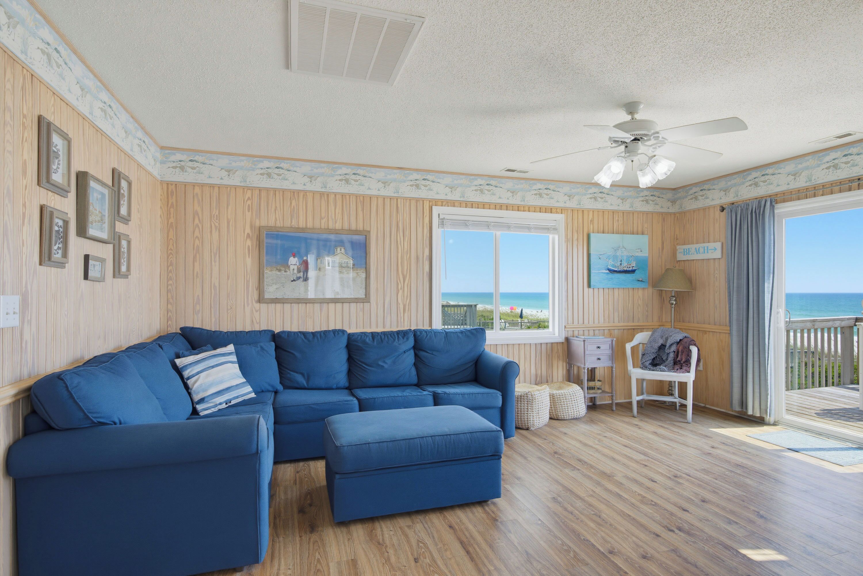 A room with a blue sectional sofa, a painting of a beach scene, a window with a view of the beach, and a ceiling fan.