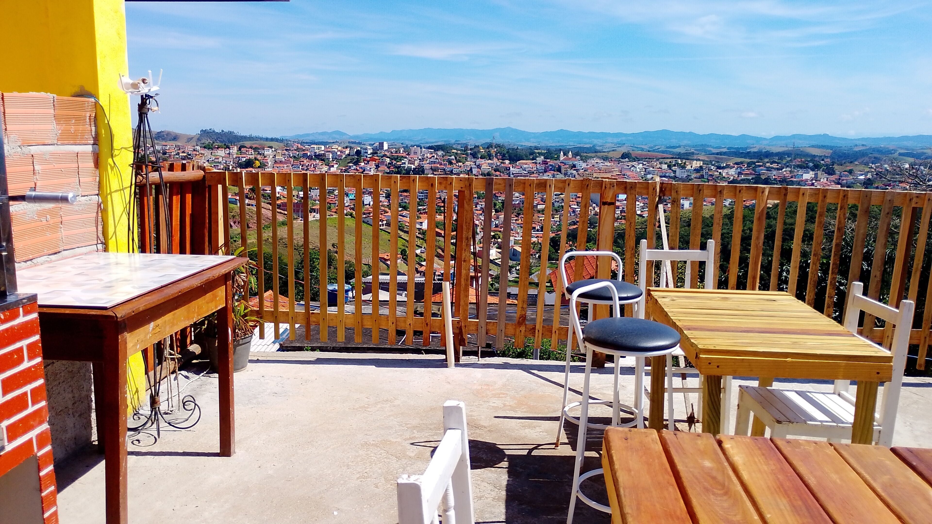 Un balcón con cercado de madera, una mesa y dos sillas, con vista a un paisaje urbano que incluye edificios y colinas.