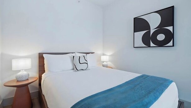 Et hotelværelse med en seng, to sengeborde med lamper, et vægmaleri i abstrakt stil og et blåt plaide.