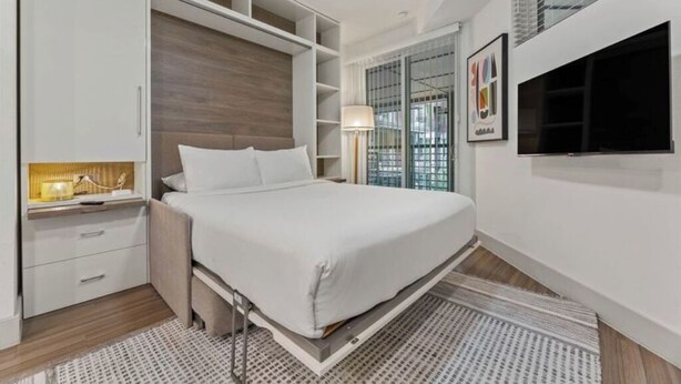 A bedroom with a bed, built-in shelving, a bedside table with a lamp, and a TV mounted on the wall.