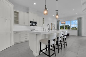 A modern kitchen with white cabinetry, a central island with bar stools, pendant lighting, and a view of the outdoors through large windows. - DOMINICA ISLES 201 | Luxury Retreat at Isles of Collier Preserve (Naples)