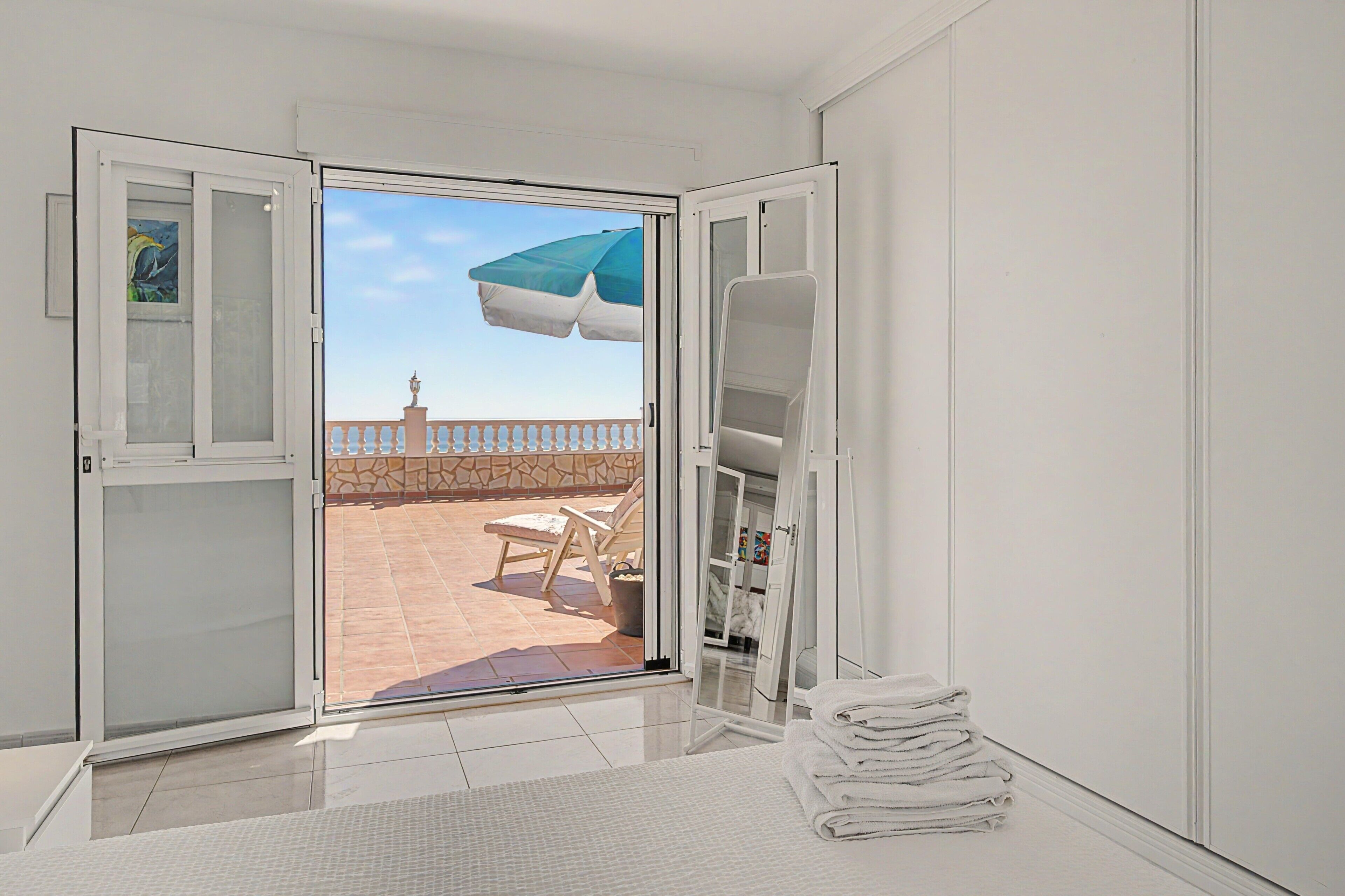 A room with a sliding glass door leading to an outdoor area with a tiled floor, a white chair, and a blue and white umbrella.