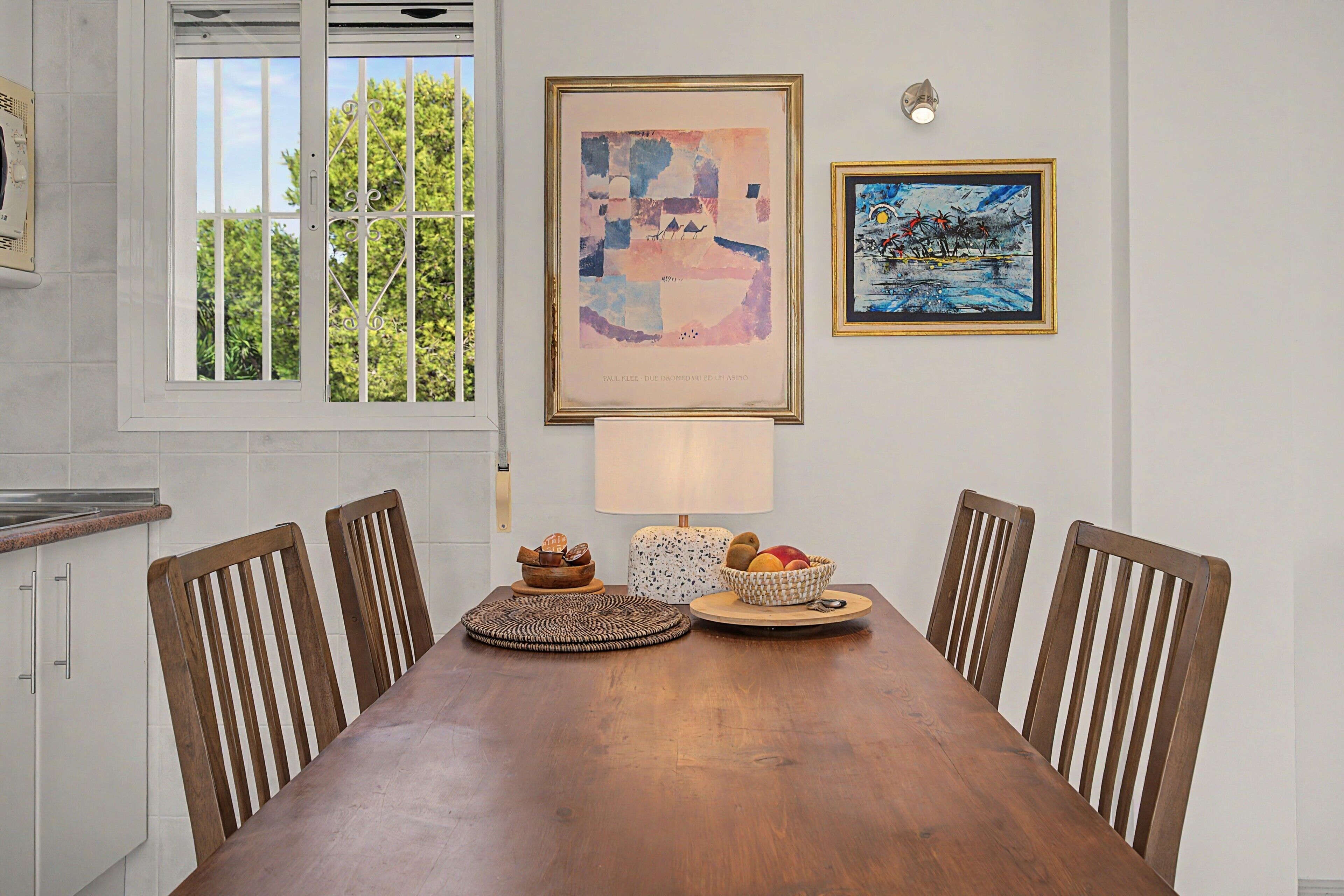 A dining area with a wooden table, six chairs, a lamp, and two framed artworks on the wall.