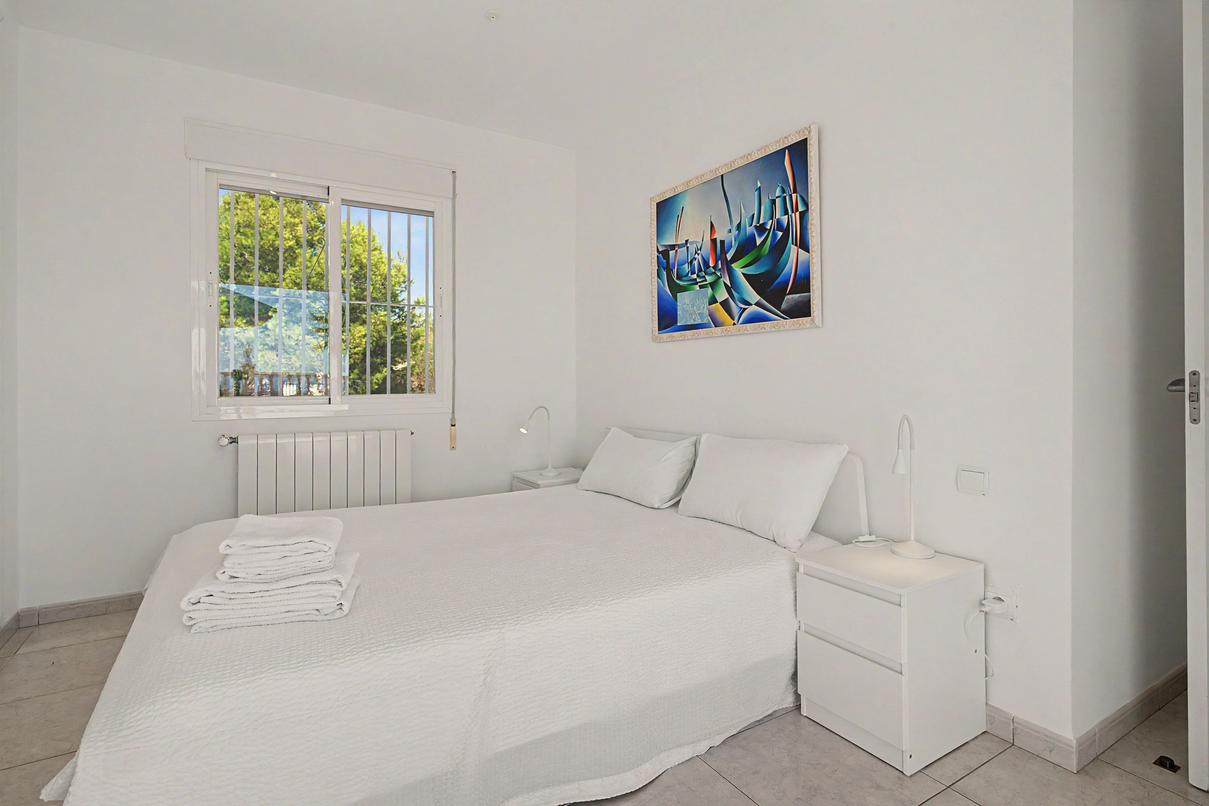A bedroom with a bed, a nightstand, a bedside lamp, a window, and a painting on the wall.