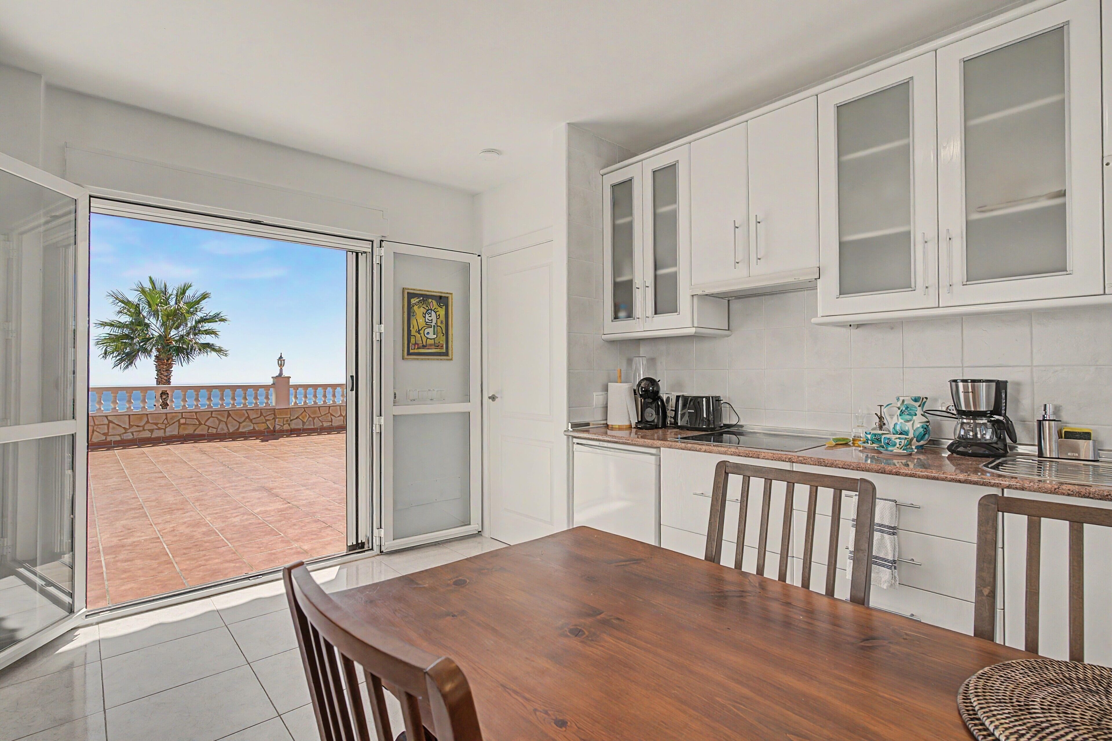 A modern kitchen with white cabinets, a wooden dining table, and a view of a palm tree and balcony.