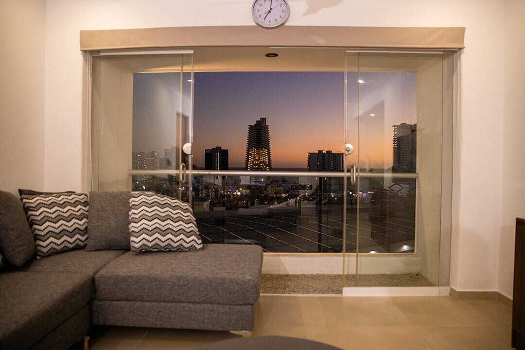 A living room with a sofa and cushions, a glass door leading to a cityscape view at dusk.