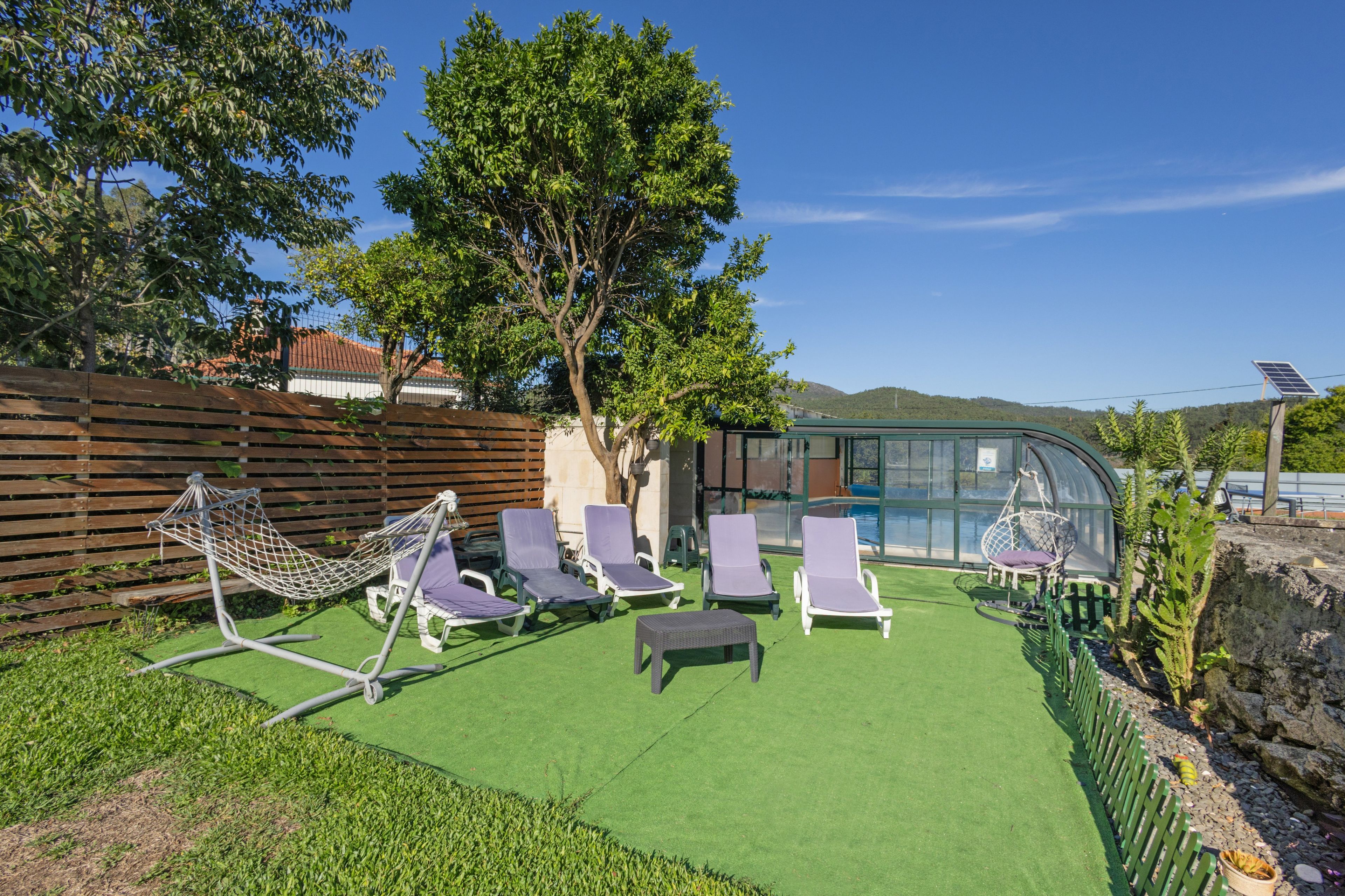 Un espacio al aire libre con sillones de descanso y una hamaca, rodeado de follaje y una estructura de vidrio.