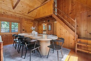 A wooden interior with a round dining table surrounded by chairs, a staircase, and large windows. - The Gingerbread House | Sauna, HotTub, Lake Access (Twain Harte)