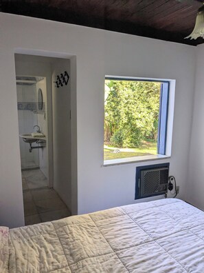 Un dormitori amb llit, finestra amb vista a vegetació, radiador i bany amb rentaplat i mirall.