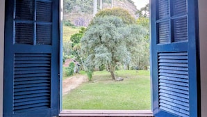 An open window with shutters, a view of a grassy area with trees, and a hill in the background.