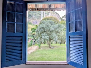 An open window with shutters, a view of a grassy area with trees, and a hill in the background.