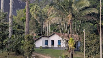 A house with a white exterior and blue shutters, surrounded by lush greenery and a mountain backdrop.