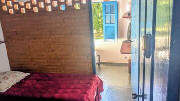 A room with a brick wall, a bed with a red blanket, and a door leading to an outdoor area with a blue shutter.