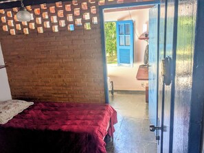 A room with a brick wall, a bed with a red blanket, and a door leading to an outdoor area with a blue shutter.