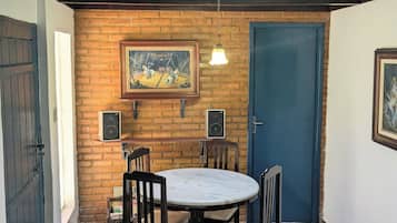 A dining area with a brick wall, a round table, four chairs, two wall-mounted speakers, and a framed picture above the table.