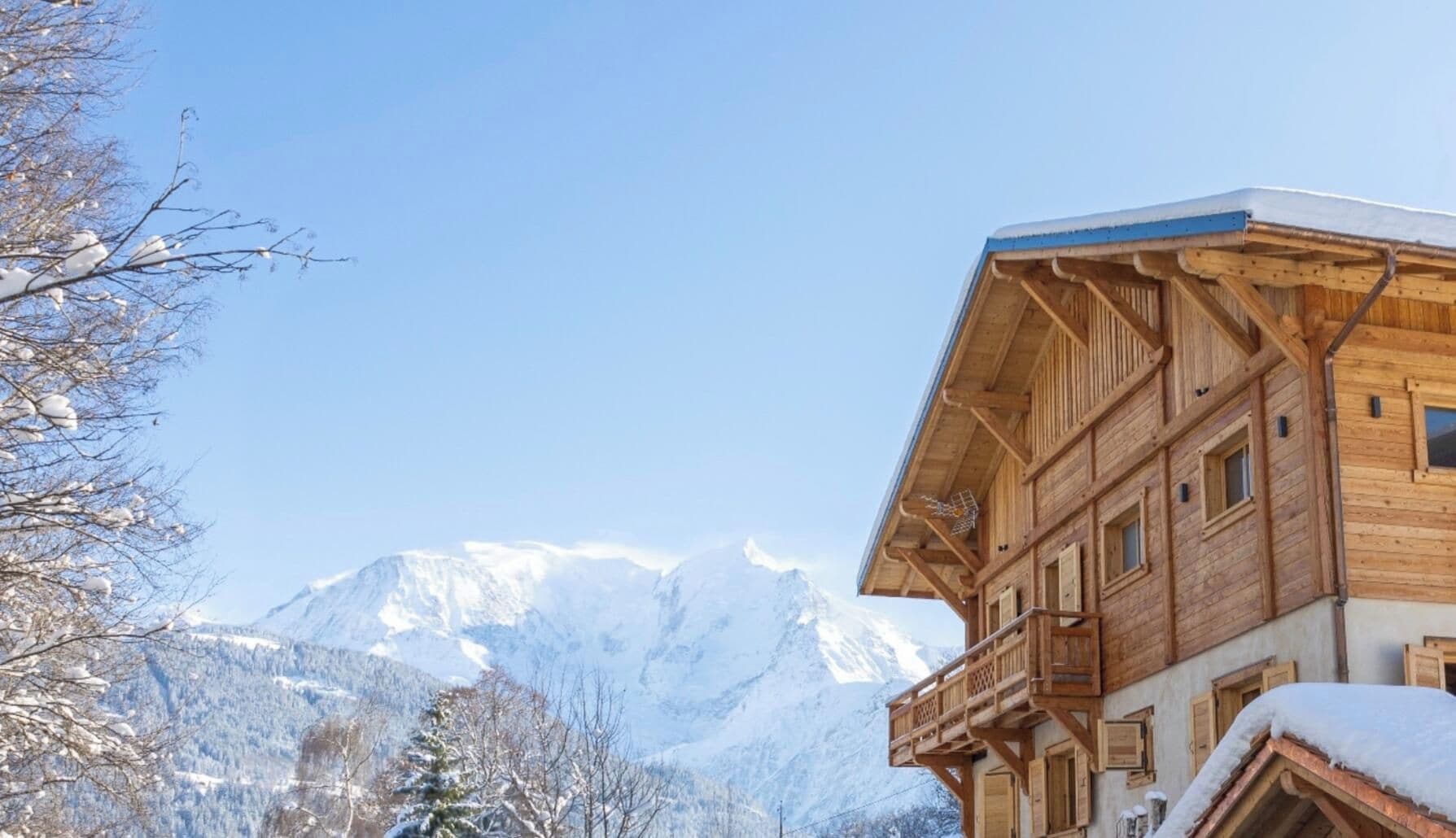 A wooden chalet with a balcony, set against a backdrop of snow-covered mountains.