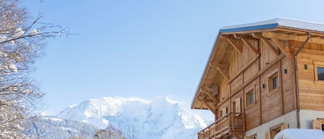 A wooden chalet with a balcony, set against a backdrop of snow-covered mountains.
