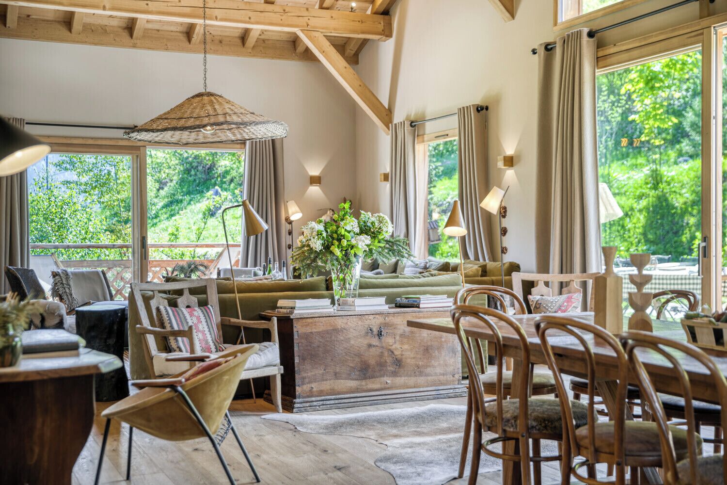A cozy room with a wooden ceiling, a sofa, chairs, a dining table, and a view of greenery outside.