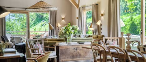 A cozy room with a wooden ceiling, a sofa, chairs, a dining table, and a view of greenery outside.