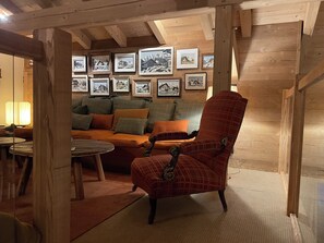Ein gemütliches Zimmer mit Holzbalkendecke, einer Couch mit Kissen, einem roten Karo-Sessel und gerahmten Bildern an der Wand.