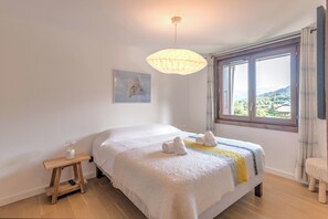 A bedroom with a bed, a bedside table, a lamp, a window with a view, and a painting on the wall. - Le Duplex des Sommets - Welkeys (Demi-Quartier)