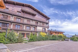 Un immeuble à plusieurs étages doté de balcons, une zone pavée à l’avant et un paysage montagneux en arrière-plan.