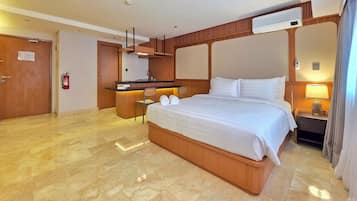 Quarto de hotel com uma cama grande, uma pequena escrivaninha, uma cadeira, um minibar e a porta do banheiro.
