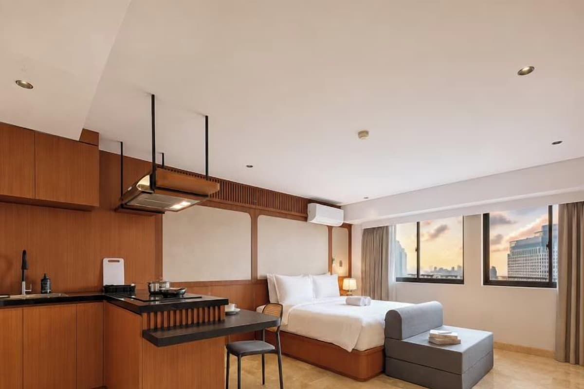 Quarto de hotel moderno com cozinha compacta, cama, área de estar e uma grande janela com vista para a cidade.