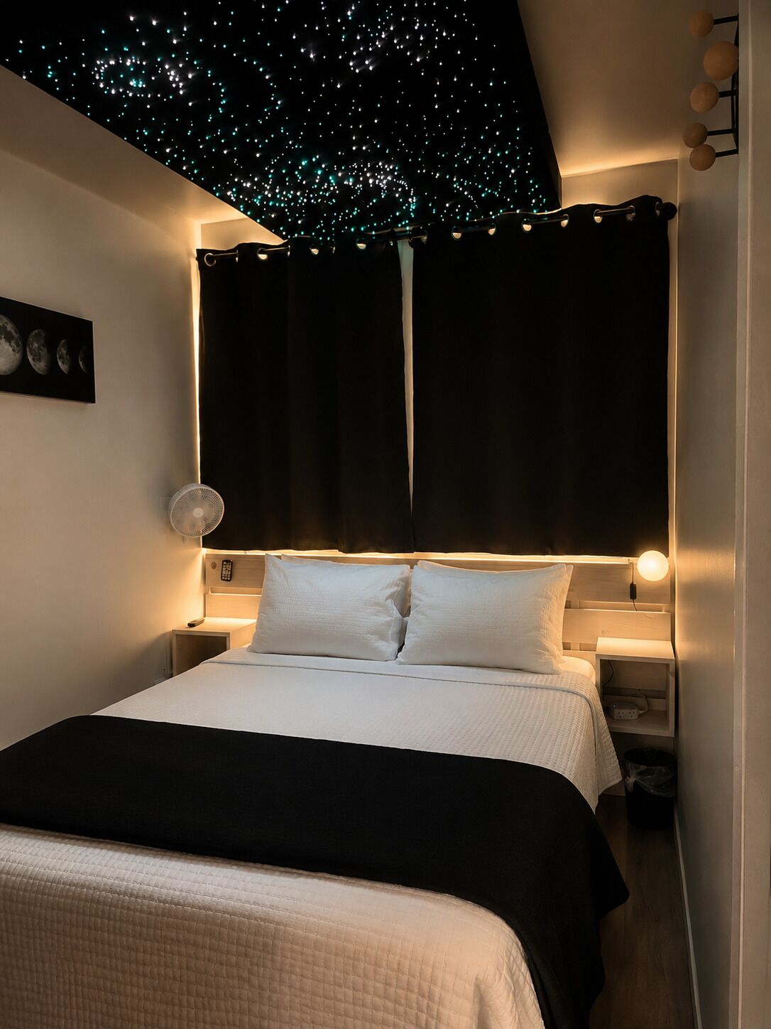 A bedroom with a bed, bedside tables, a wall-mounted light, a ceiling with star-like lights, and a partial view of a nightstand.