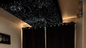 Un dormitorio con cama, mesitas de noche, una lámpara de pared, un techo con luces tipo estrellas y una vista parcial de una mesita de noche.