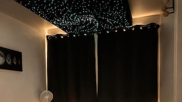 A bedroom with a bed, bedside tables, a wall-mounted light, a ceiling with star-like lights, and a partial view of a nightstand.