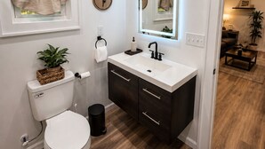 Un baño moderno con inodoro blanco, muebles de madera oscura, lavamanos blanco y un espejo con luz superior.