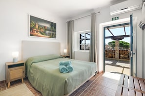 A hotel room with a bed, a nightstand, a lamp, a painting, a window, and an air conditioning unit.