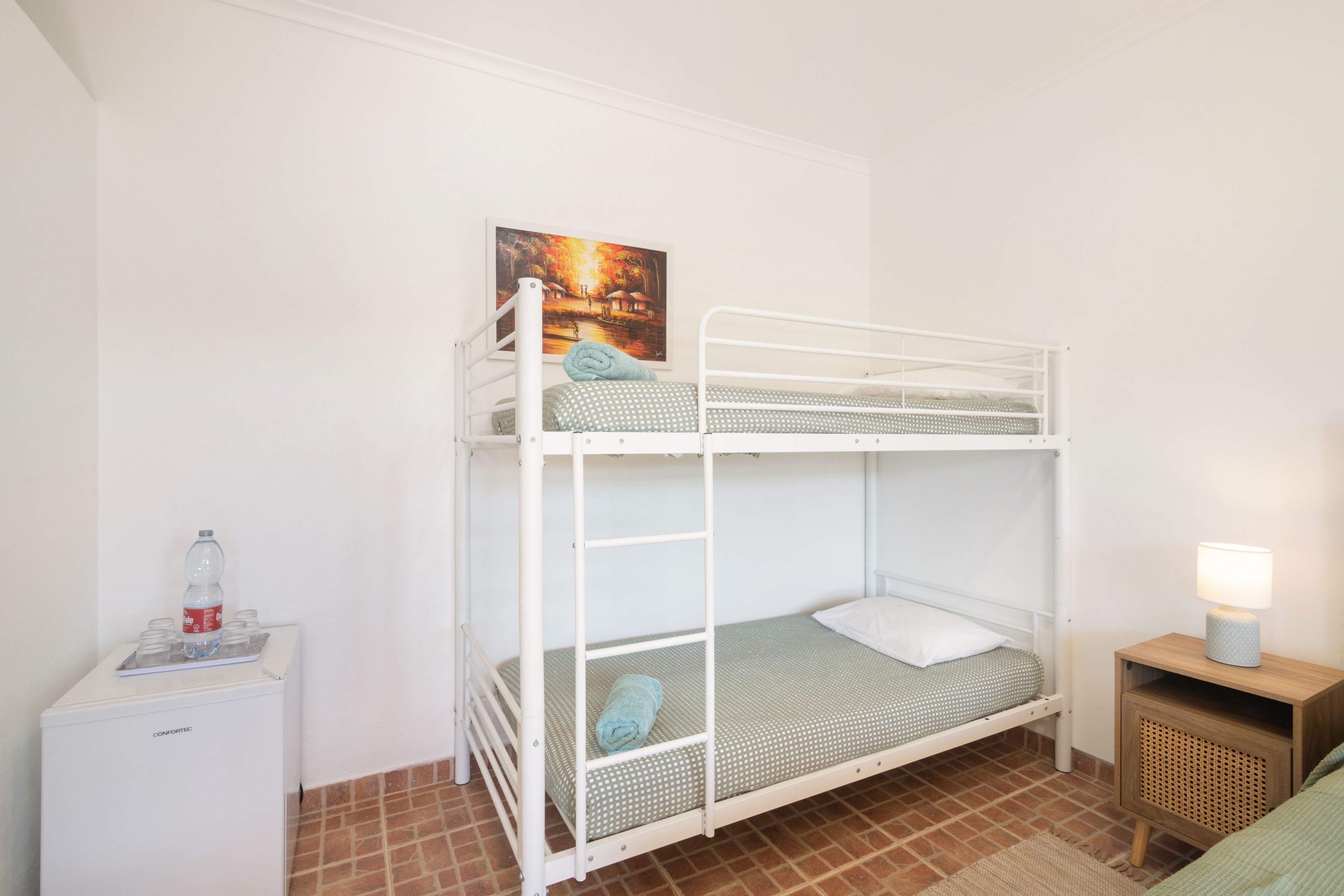 Una cama litera con un almohadón y una toalla enrollada, una mesita de noche con una lámpara y un cuadro en la pared.